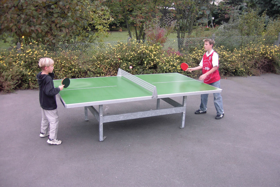 Two boys play table tennis on a blue Butterfly S2000 Polymer Concrete/Steel table with square corners. Surrounded by trees, both hold paddles and appear focused. The sturdy Butterfly table is designed for permanent outdoor use.