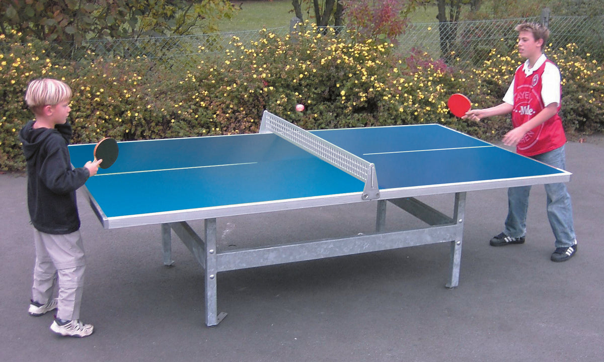 Two boys play table tennis outside on a blue Butterfly S2000 polymer concrete/steel table with rounded corners. One wears a black jacket, the other a red and white shirt. Yellow flowers and greenery are visible in the background.