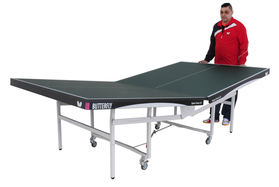 A man in a red and black jacket stands next to a green Butterfly Space Saver 22 Rollaway Table Tennis Table, which has one half upright for solo play or storage and is wheelchair-friendly.
