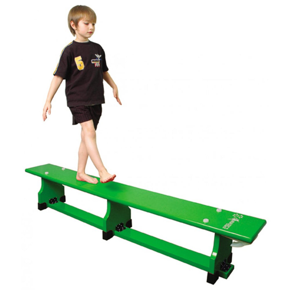 A young child in a black sports outfit walks barefoot, arms out, on a green Sure Shot Coloured Balance Bench (11ft) by Sure Shot, set against a white background.