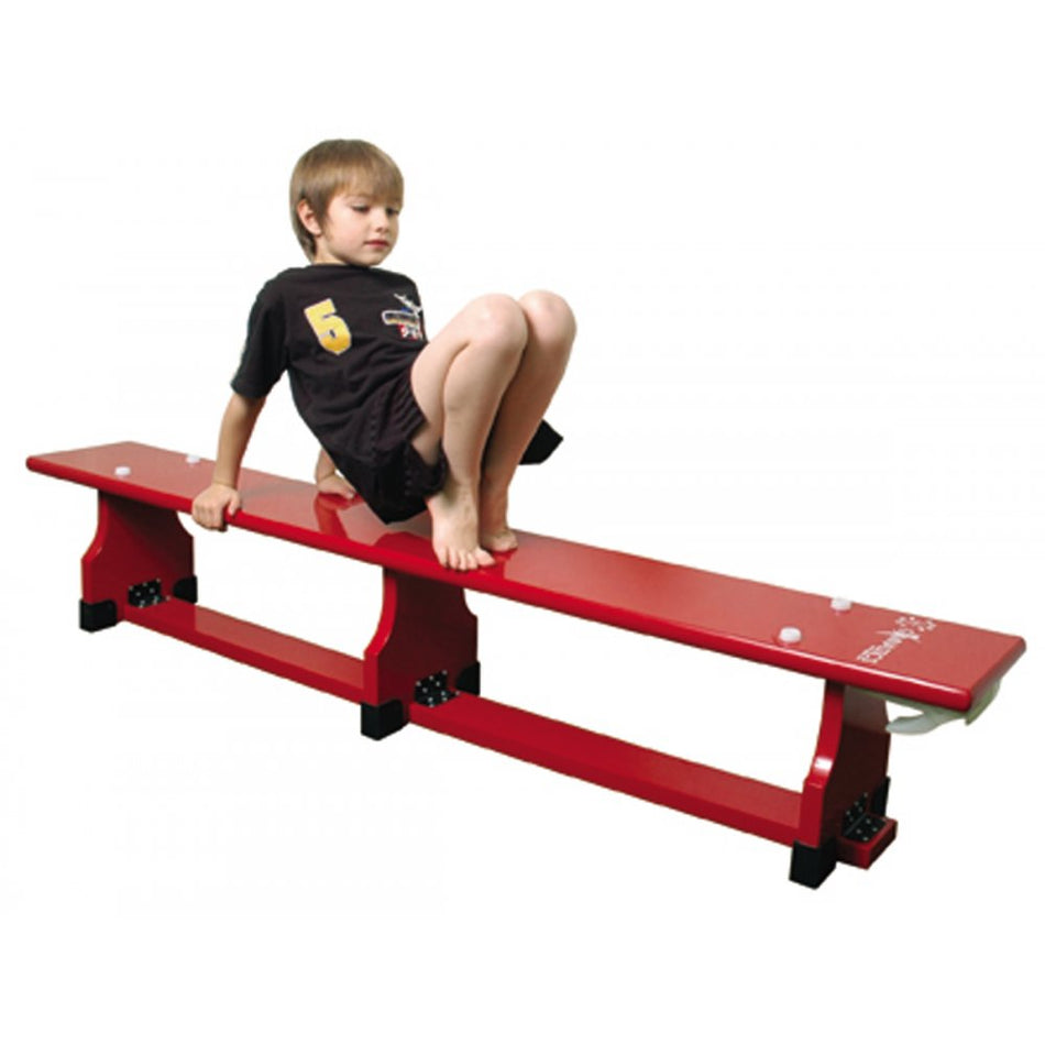 A young boy in a black shirt and shorts balances barefoot on a Sure Shot Coloured Balance Bench (6ft) by Sure Shot, using his hands and feet to steady himself as he moves along the red, knotless wood bench.