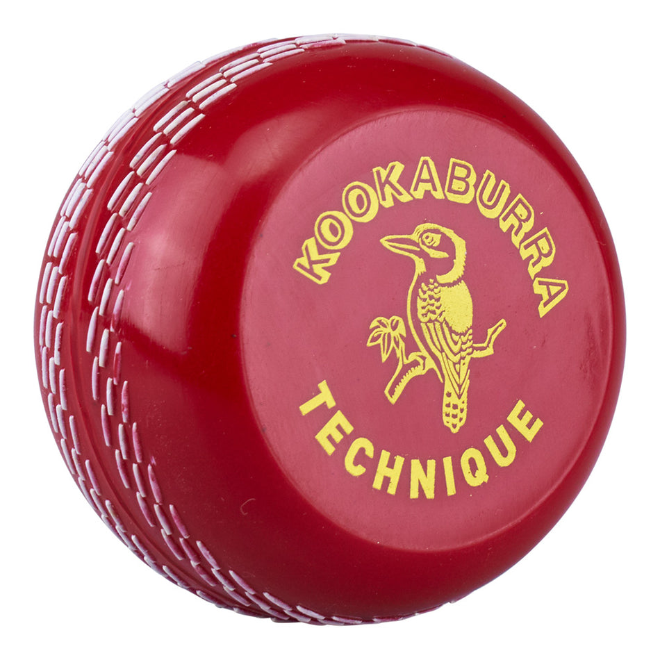 The Kookaburra Technique Trainer is a red cricket ball with white stitching, featuring the yellow Kookaburra logo—a bird on a branch—and “KOOKABURRA TECHNIQUE TRAINER” text to help improve your release skills.