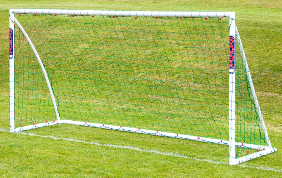The Samba Sports Trainer Goal 16 x 7, a white freestanding football goal with green netting and a secure locking system, stands on grass marked with white field lines. The "SAMBA" logo is visible on the side posts.