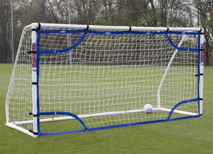 The Samba Sports Target net 12 x 6 portable football goal in white and blue is set up on a grassy field, with a soccer ball near the back of the net. Ideal for shooting drills, with trees and a fence in the background.