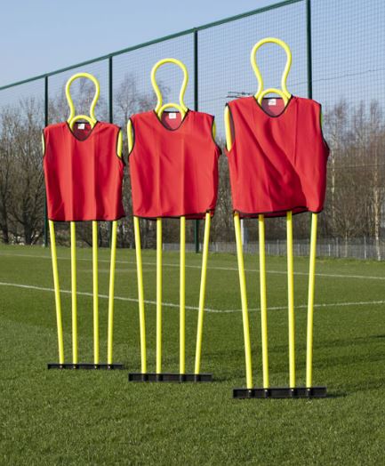 Three Samba Sports Pep Pro Mannequins (set of 3, with carry bag) in red vests stand on a green soccer field by a wire fence—ideal for free kick training or use as coaching mannequins during soccer practice.
