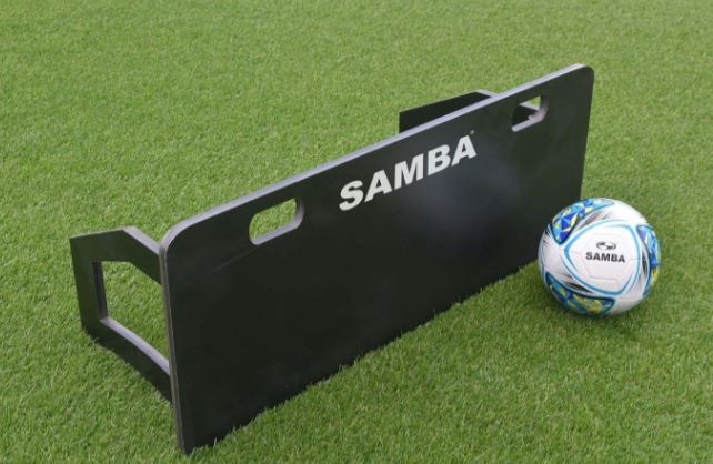 A Samba Sports Samba Rebound Board in black rests on green artificial grass beside a blue and white soccer ball, perfect for enhancing passing accuracy and ball control.