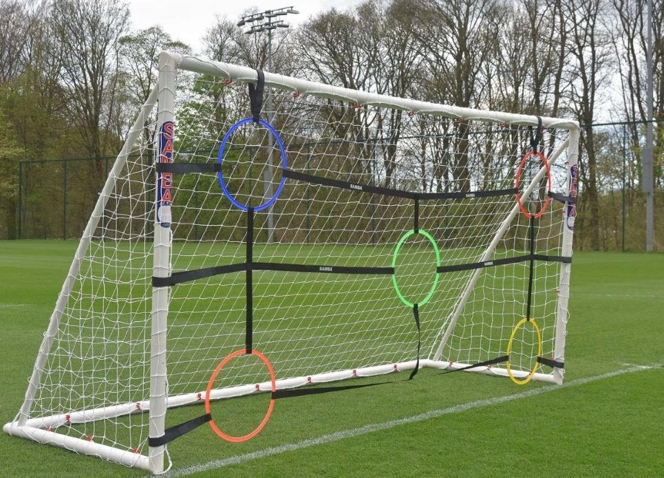 The Samba Sports Target Net 12 x 6 with hoops features six colored rings—two on each side, one top center, and one bottom center—for shooting accuracy training on a grass field. Trees and lights in the background create an ideal training environment.