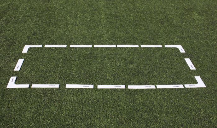 A Samba Sports Samba Pitch Marker Set creates a rectangular outline with white rubber markers on green grass, marking designated coaching areas. Some markers are labeled, but the wording is unclear.