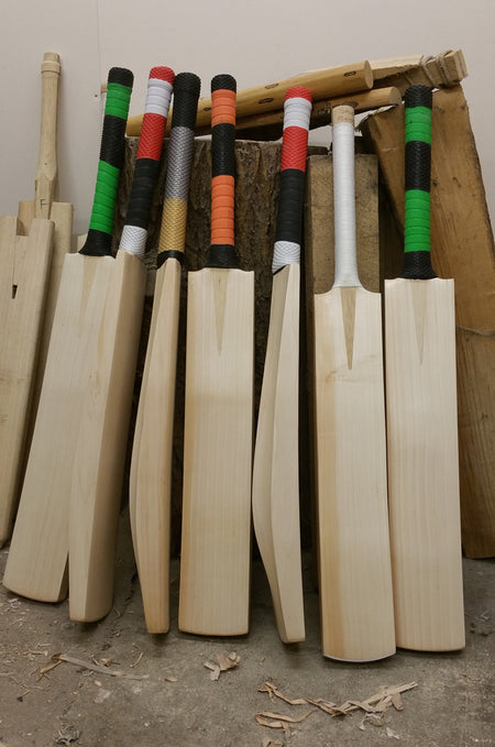 Seven Martin Berrill Sports Custom Made Cricket Bats (Grade 1+ Willow) – Pro Players Edition, featuring colorful grips, stand upright in a workshop amid wood shavings and unfinished wooden pieces.