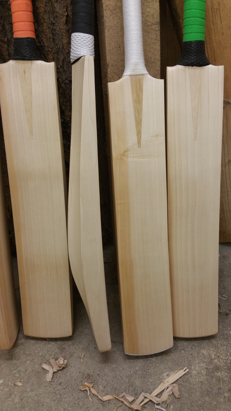 Four Martin Berrill Sports Custom Made Cricket Bat (Grade 1+ Willow) - Pro Players Edition stand upright with varied colored grips on a workshop floor, one turned sideways to display its profile amid scattered wood shavings.