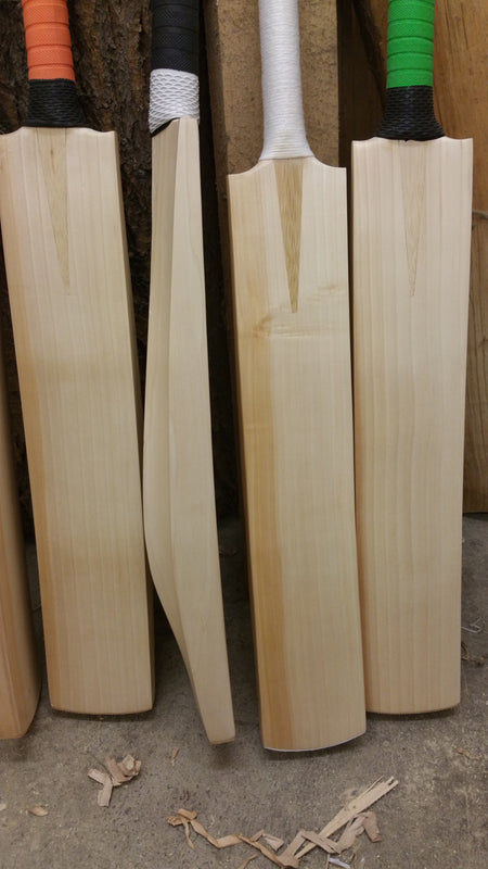 Four Martin Berrill Sports Custom Made Cricket Bat (Grade 1+ Willow) - Pro Players Edition stand upright with varied colored grips on a workshop floor, one turned sideways to display its profile amid scattered wood shavings.