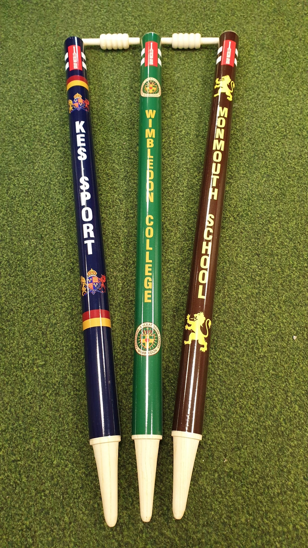 Three Custom Printed Club Cricket Stumps by Gray Nicolls, featuring pointed ends and custom labels, are displayed upright on green turf with a wooden bail resting on top.