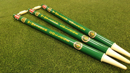 Three Gray Nicolls Custom Printed Club Cricket Stumps with green bodies, white ends, yellow text, and logos are arranged on artificial grass. The bails rest on top of two stumps.