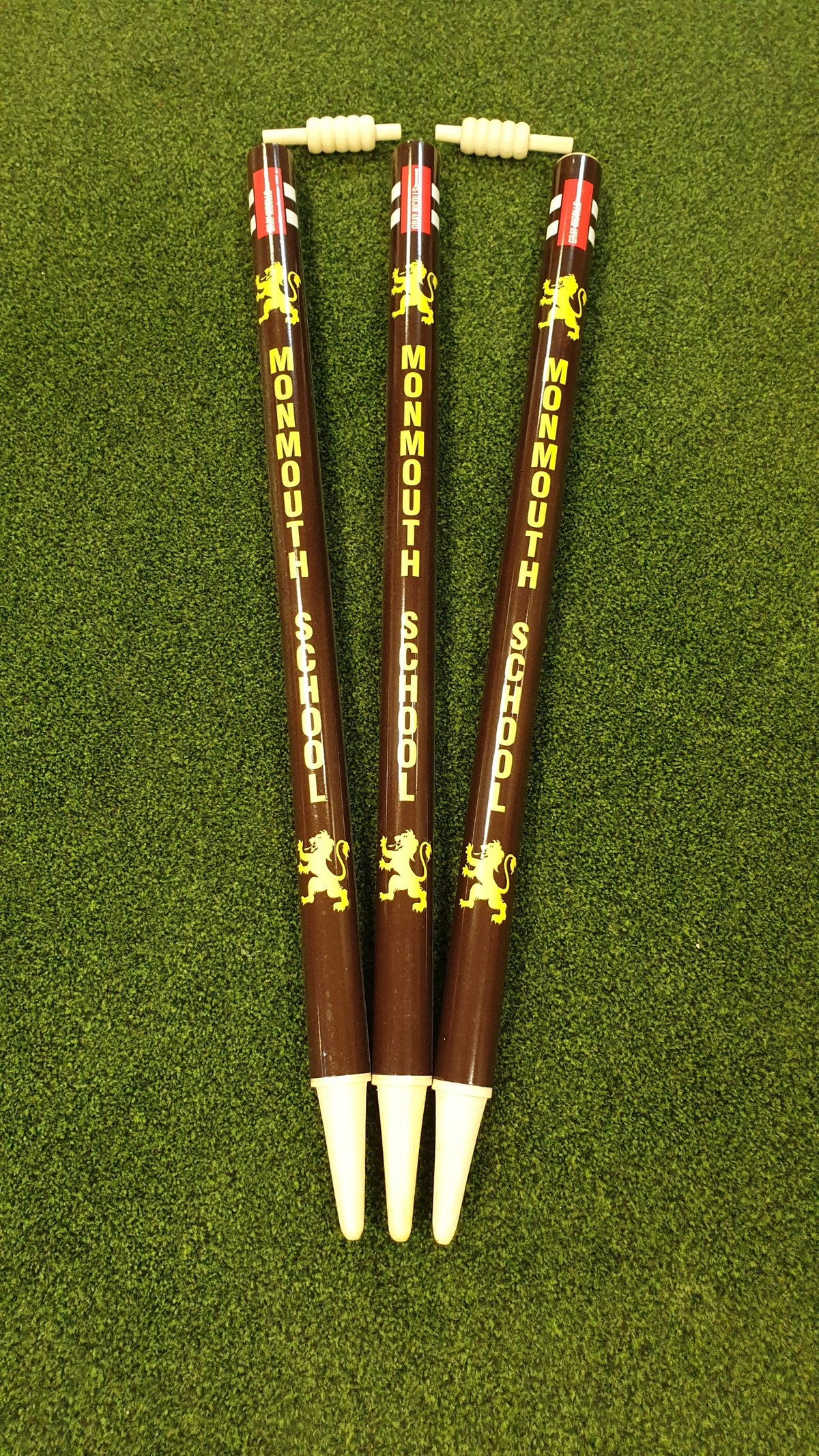 Three brown ballpoint pens, inspired by Gray Nicolls Custom Printed Club Cricket Stumps, display Monmouth School and a yellow lion emblem. Each pen stands tip-down on green fabric and features a small abacus at the top.