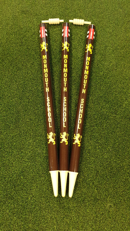 Three brown ballpoint pens, inspired by Gray Nicolls Custom Printed Club Cricket Stumps, display Monmouth School and a yellow lion emblem. Each pen stands tip-down on green fabric and features a small abacus at the top.