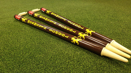 Three brown wooden Custom Printed Club Cricket Stumps by Gray Nicolls, with white ends and yellow branding featuring a lion logo, are laid parallel on green synthetic grass.