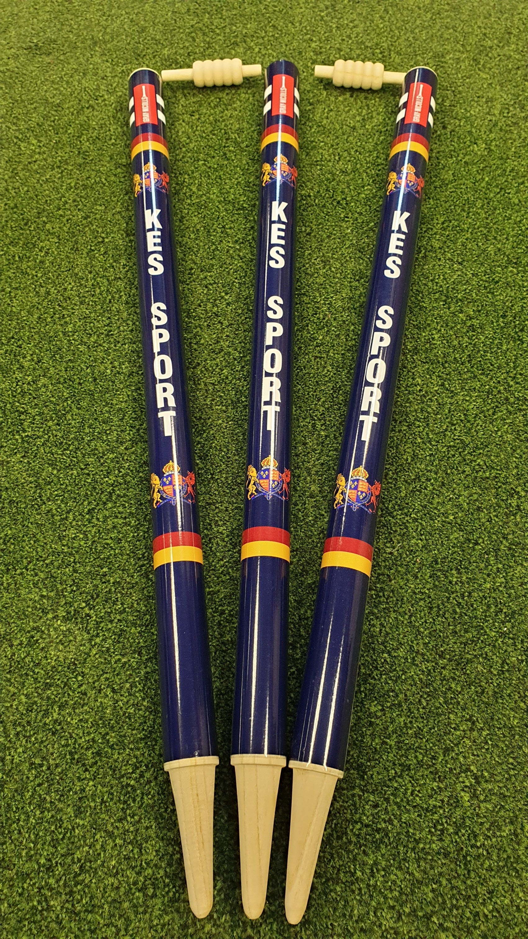 Three blue Custom Printed Club Cricket Stumps by Gray Nicolls with beige bases stand upright on green artificial turf, while a disassembled beige bail rests above the stumps.