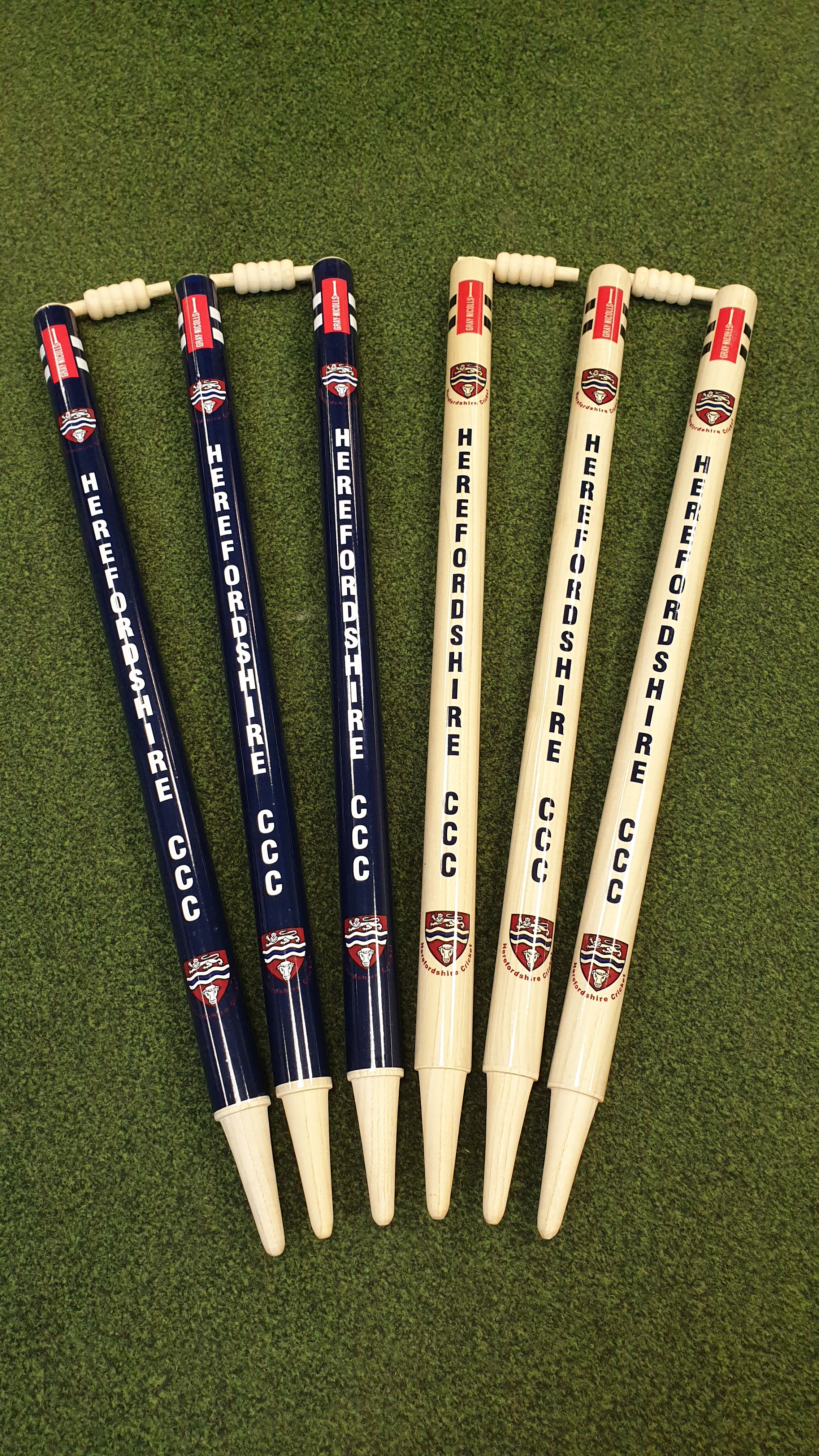 Six Gray Nicolls Custom Printed Club Cricket Stumps—three dark blue, three light wood—all with HEREFORDSHIRE CCC and a crest, stand on green artificial grass with two bails on top.