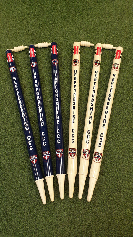 Six Gray Nicolls Custom Printed Club Cricket Stumps—three dark blue, three light wood—all with HEREFORDSHIRE CCC and a crest, stand on green artificial grass with two bails on top.