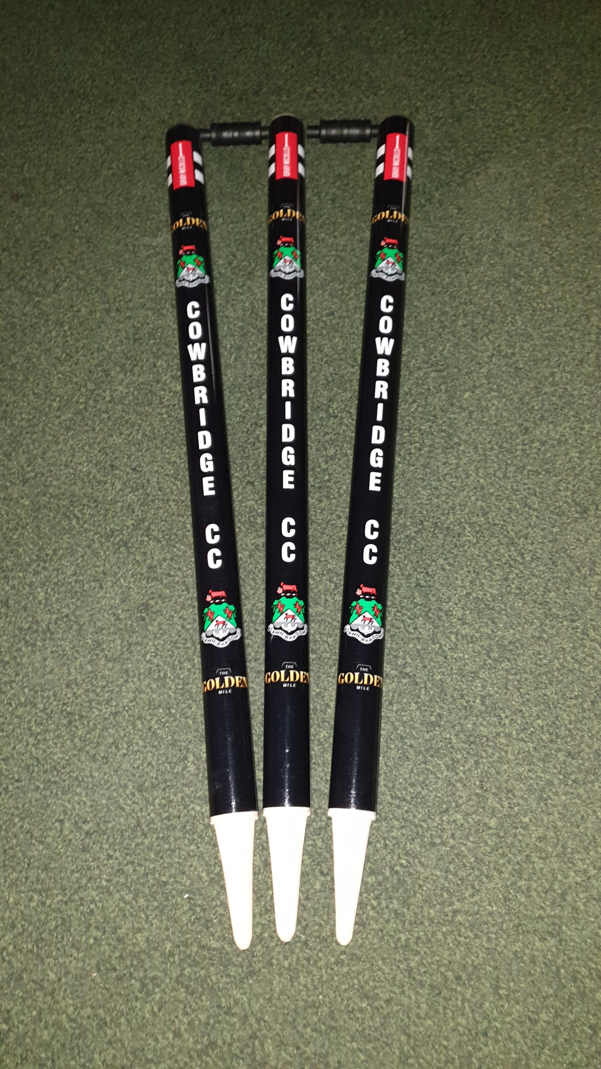 Three black Gray Nicolls Custom Printed Club Cricket Stumps featuring COWBRIDGE CC and colorful logos stand upright on green turf, with red labels on top and white bases—ideal personalised stumps for any cricket match.