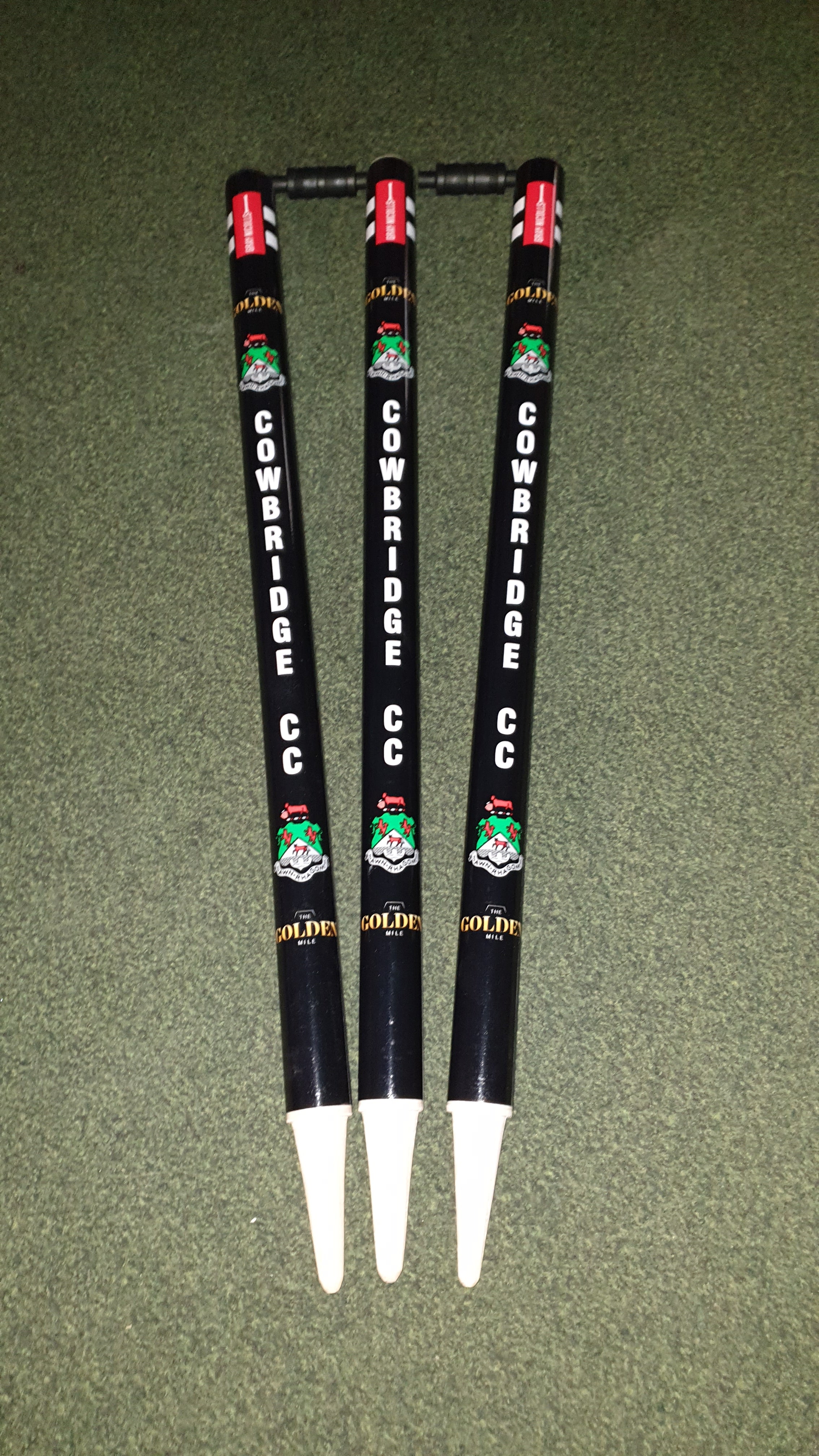 Three black Gray Nicolls Custom Printed Club Cricket Stumps featuring COWBRIDGE CC and colorful logos stand upright on green turf, with red labels on top and white bases—ideal personalised stumps for any cricket match.