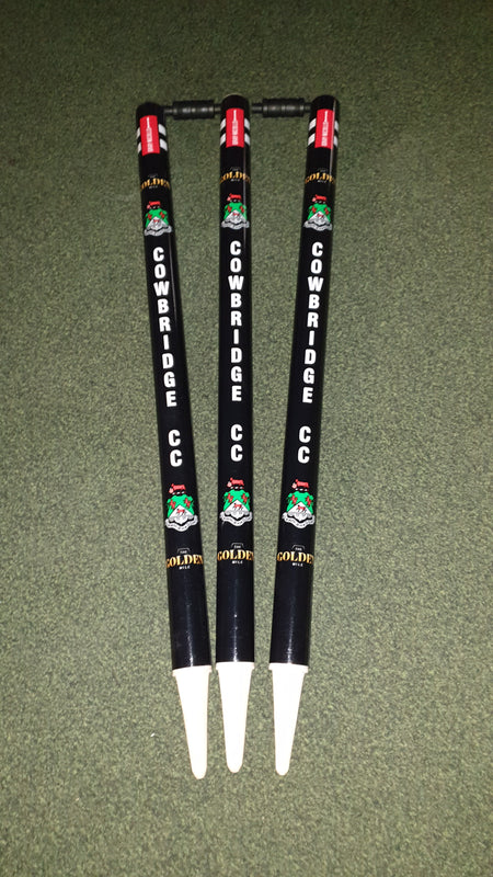 Three black Gray Nicolls Custom Printed Club Cricket Stumps featuring COWBRIDGE CC and colorful logos stand upright on green turf, with red labels on top and white bases—ideal personalised stumps for any cricket match.