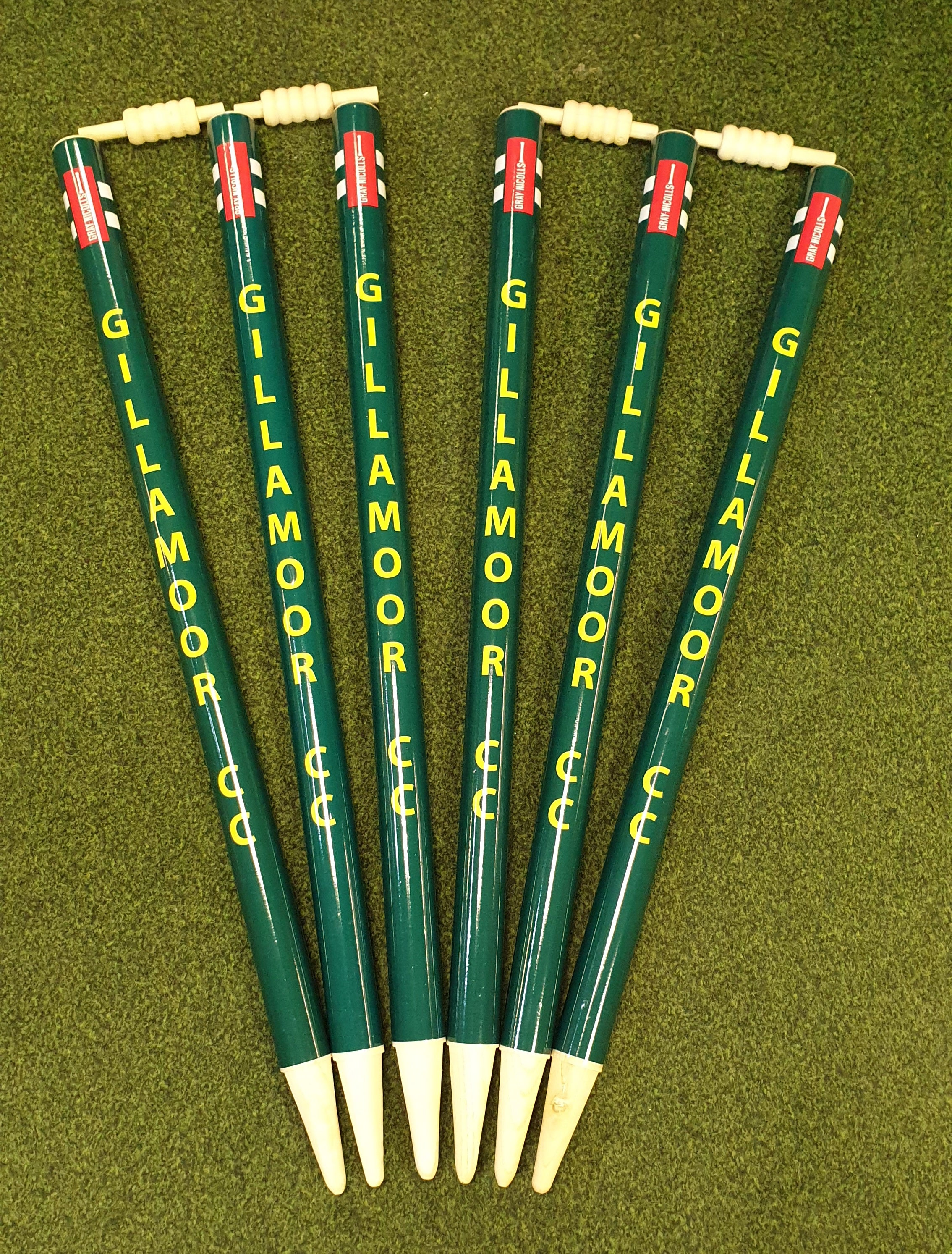 Five green Gray Nicolls Custom Printed Club Cricket Stumps with yellow GILLAMOOR CC text are arranged fan-shaped on grass, each personalised stump topped with a bail.