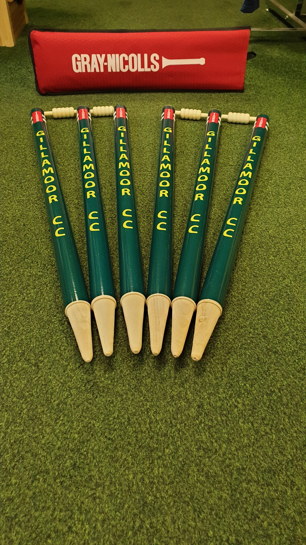 Six green Custom Printed Club Cricket Stumps by Gray Nicolls, printed with KILMOR CC and arranged in a fan shape on green turf with wooden bails on top, are shown with a red GRAY-NICOLLS bag in the background.