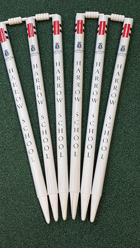 Six white Gray Nicolls Custom Printed Club Cricket Stumps, each labeled HARROW SCHOOL in blue, fan-shaped on green. The tops feature mini abacuses and bat handles, resembling personalized stumps for schools and clubs.