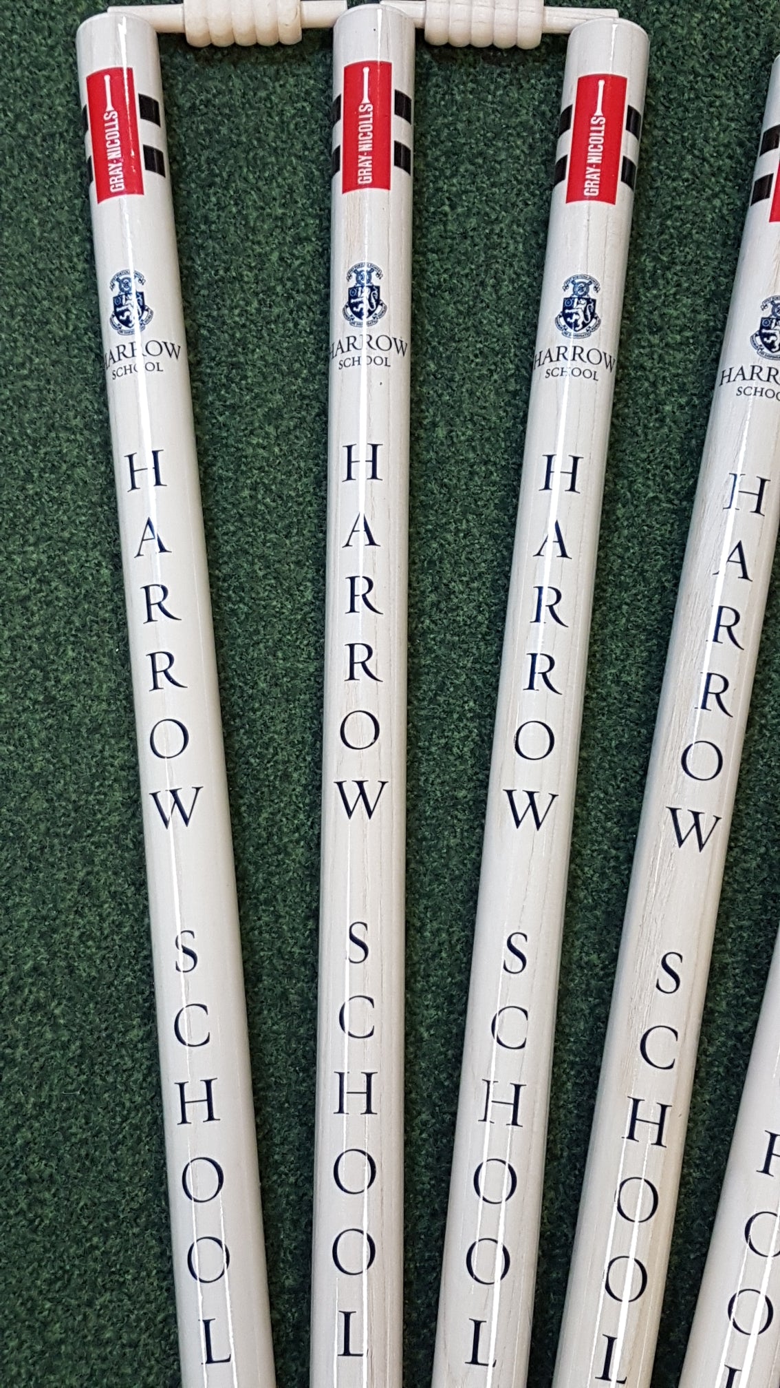 Five Custom Printed Club Cricket Stumps by Gray Nicolls, white with red bails on green turf, each stump vertically labeled with Harrow School and a crest logo.