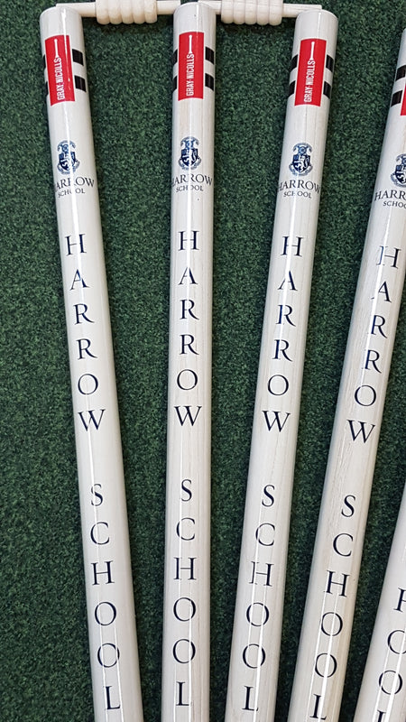 Five Custom Printed Club Cricket Stumps by Gray Nicolls, white with red bails on green turf, each stump vertically labeled with Harrow School and a crest logo.