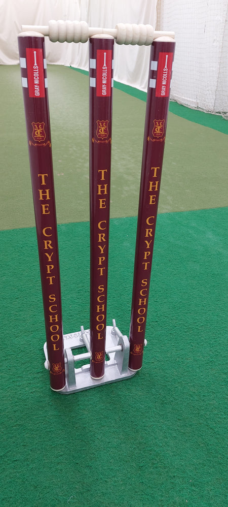 Three maroon Gray Nicolls Custom Printed Spring Back Stumps, printed with The Crypt School, set up on a green indoor pitch with white bails—perfect for cricket practice sessions.