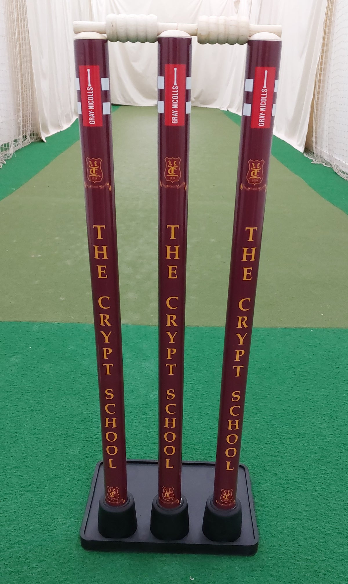 Three maroon Gray Nicolls Custom Printed Match Cricket Stumps with a heavy rubber base, featuring "THE CRYPT SCHOOL" in yellow vertically, set on a green indoor pitch with white side netting.