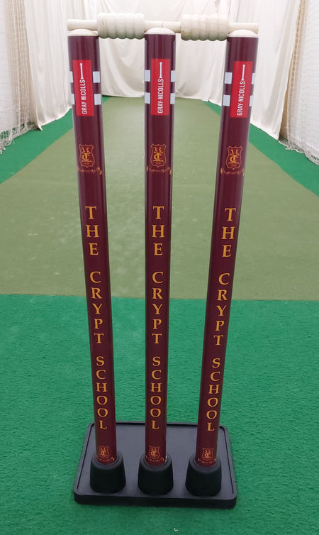 Three maroon Gray Nicolls Custom Printed Match Cricket Stumps with a heavy rubber base, featuring "THE CRYPT SCHOOL" in yellow vertically, set on a green indoor pitch with white side netting.