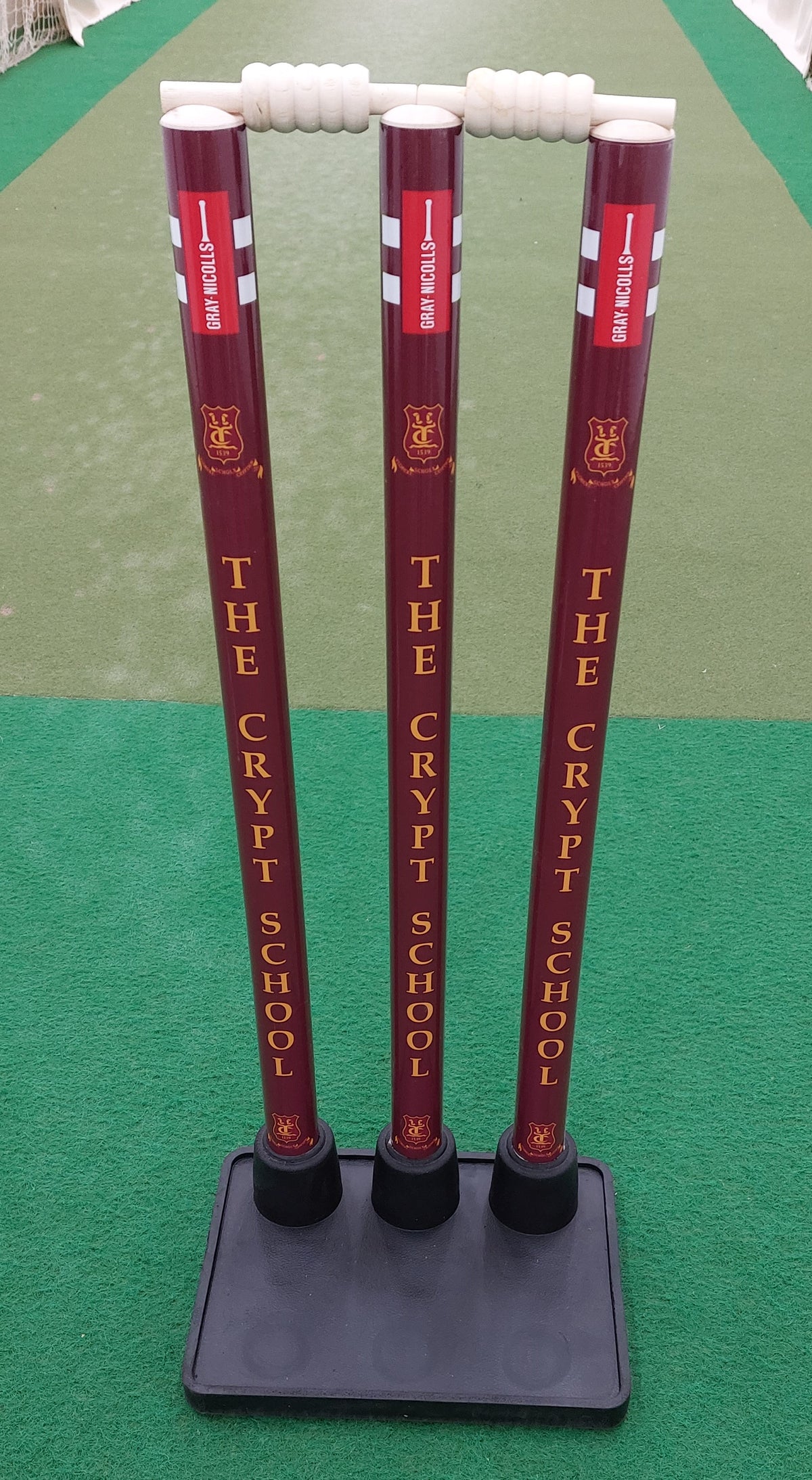 Three maroon Gray Nicolls Custom Printed Match Cricket Stumps with heavy rubber base and white bails, featuring THE CRYPT SCHOOL and a lion crest, are set on a green artificial cricket pitch.