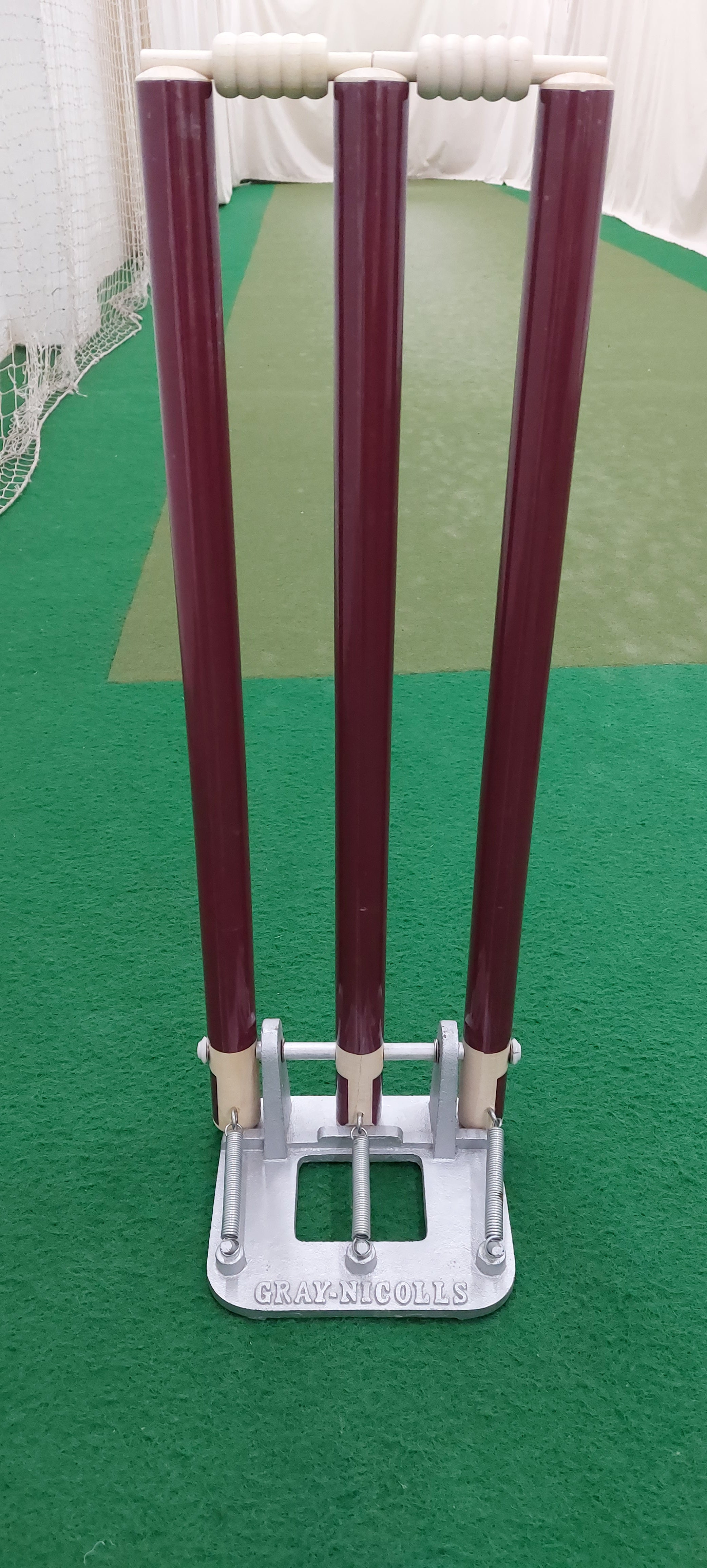 The Gray Nicolls Custom Printed Spring Back Stumps are a set of three maroon cricket stumps with white bails on a metallic base, perfect for training and placed on green indoor practice turf.