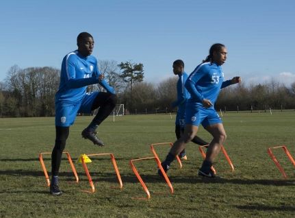 Three soccer players in blue train outdoors, jumping over Samba Sports Samba Speed and Agility Hurdles on a grassy field with trees and a clear sky in the background.