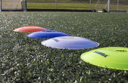 Four Samba Flat Marker cones by Samba Sports—in orange, blue, white, and yellow—are arranged on artificial grass beside soccer goals and nets; perfect coaching markers for drills.