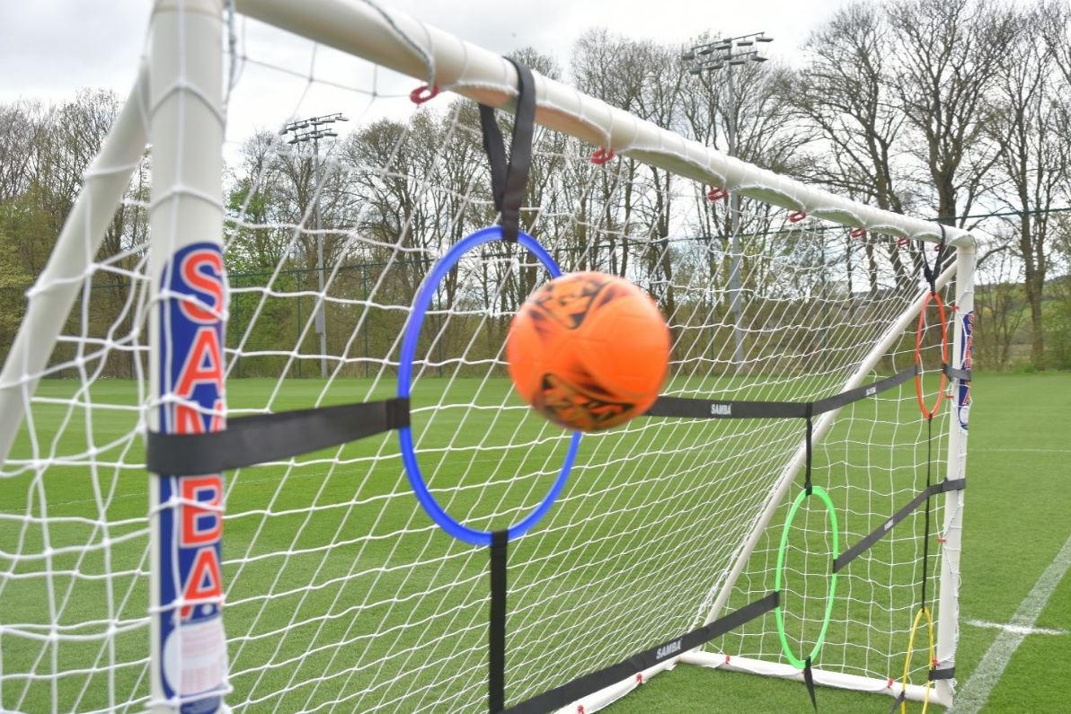 An orange soccer ball passes through a blue hoop on the Samba Sports Target net 12 x 6 with hoops, set on a green field. Green and yellow hoops also hang from this training tool, with trees and floodlights in the background.