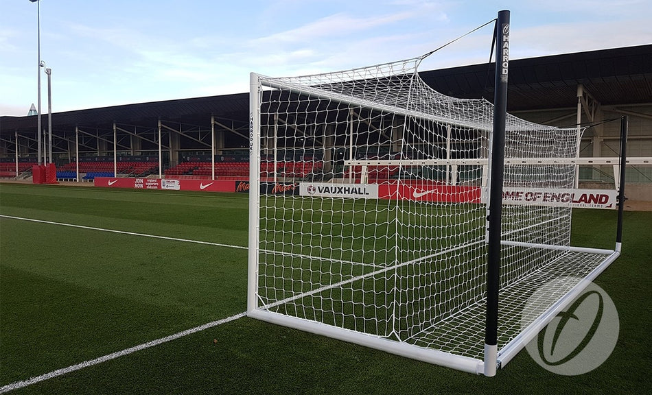 A Harrod Sport 4G UEFA Portagoal, crafted from reinforced aluminium, stands on green artificial turf near an empty stadium lined with red seats and advertising banners along the sideline, showcasing the premium quality of this professional-grade goal.