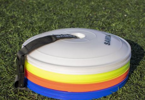 The Samba Sports Samba Flat Marker set, featuring white, yellow, orange, and blue cones with a black carrying strap, sits on green artificial grass—ideal as coaching markers or for marking fields during sports practice.