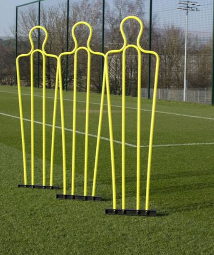 Three yellow Samba Sports Pep Pro Mannequins—with carry bag (set of 3)—are set up near the penalty area on a green soccer field, perfect for free kick training, with a wire fence and trees in the background.