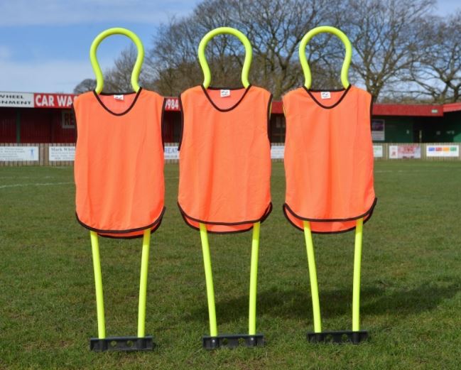 Three Samba Sports Pep Pro Junior Mannequins (set of 3, includes carry bag) stand on a grassy field wearing orange vests, ideal for free kick practice, with trees and red buildings in the background.