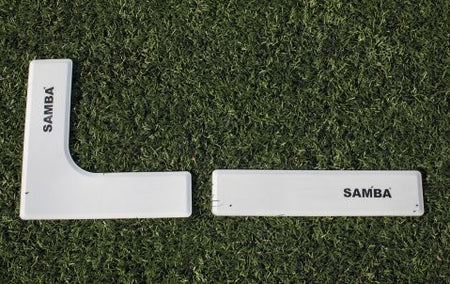 The Samba Pitch Marker Set by Samba Sports includes two white plastic markers—one L-shaped and one rectangular—positioned on artificial grass, ideal for marking coaching areas on the field.