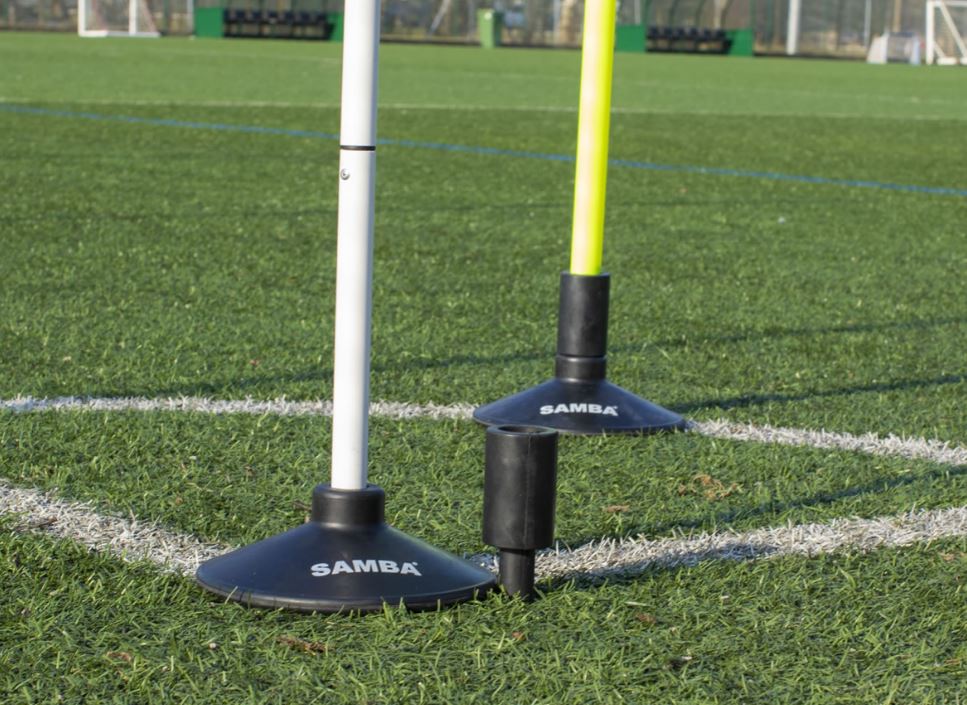 Two corner flag poles, one white and one yellow, stand upright in Samba Sports Multi-Purpose Heavy Duty Pole Bases on a grass soccer field near a white corner line. A small black attachment lies in front of the bases.