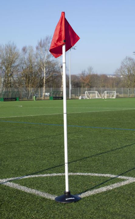 A Multi-Purpose Heavy Duty Pole Base from Samba Sports supports a red corner flag at the edge of a green artificial turf soccer field, with goals, trees, and a clear sky in the background.