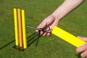 A person prepares to set up the Fusion Multi Stump Package by Fusion, holding a yellow cricket stump and a black metal spike on grass; in the background, three yellow stumps stand upright.
