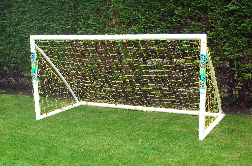 The Samba Sports Trainer Goal 8 x 4, a white uPVC football goal with yellow netting and "Samba" printed on the posts, is set up on a grassy lawn with tall green hedges—ideal for garden football games.