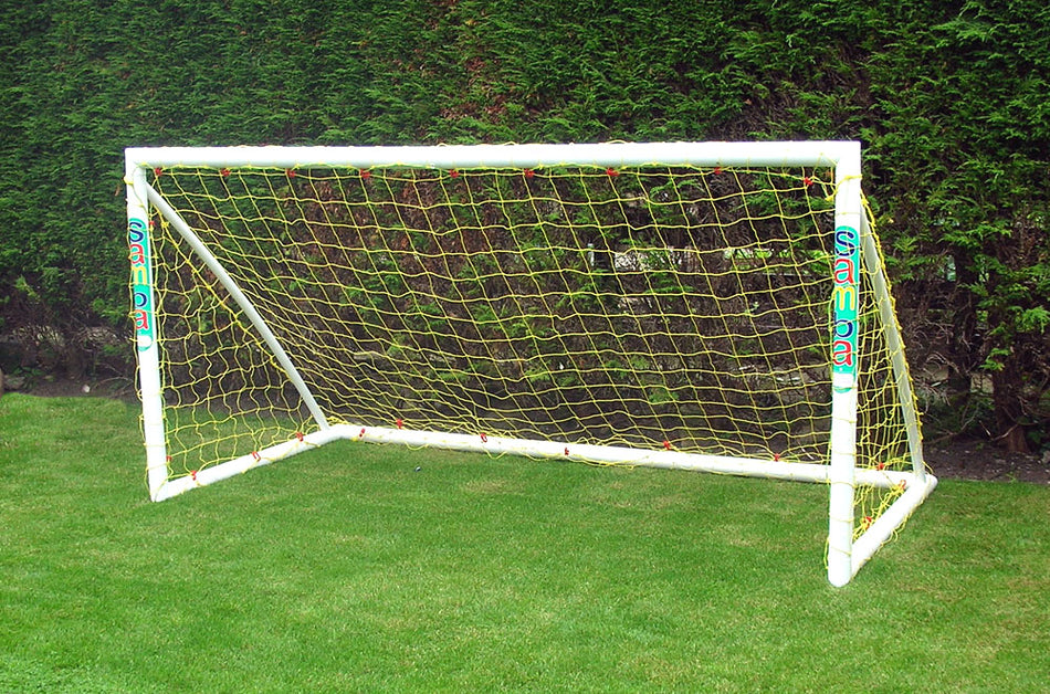 The Samba Sports Trainer Goal 8 x 4, a white uPVC football goal with yellow netting and "Samba" printed on the posts, is set up on a grassy lawn with tall green hedges—ideal for garden football games.