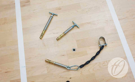 Three Harrod Sport SH2 Sportshall Anchors for solid floors (160mm) and a braided metal cable are positioned on a light wooden gym floor with white lines, centered around a small hole near recessed floor sockets.